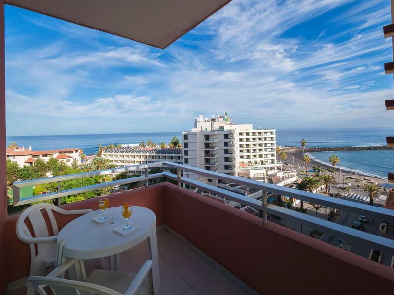 Balkon mit weißem Tisch, zwei Stühlen, Blick auf beleuchtete Gebäude und blauen Himmel mit Wolken.