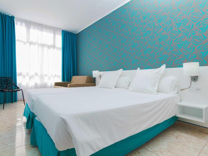 Schlichtes Schlafzimmer mit großem Doppelbett, türkisfarbenen Wänden und weißen Vorhängen.