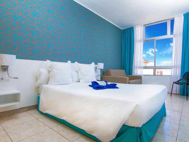 Helles Hotelzimmer mit Doppelbett, weißen Kissen, türkisfarbenen Vorhängen und Fenster mit Meerblick.