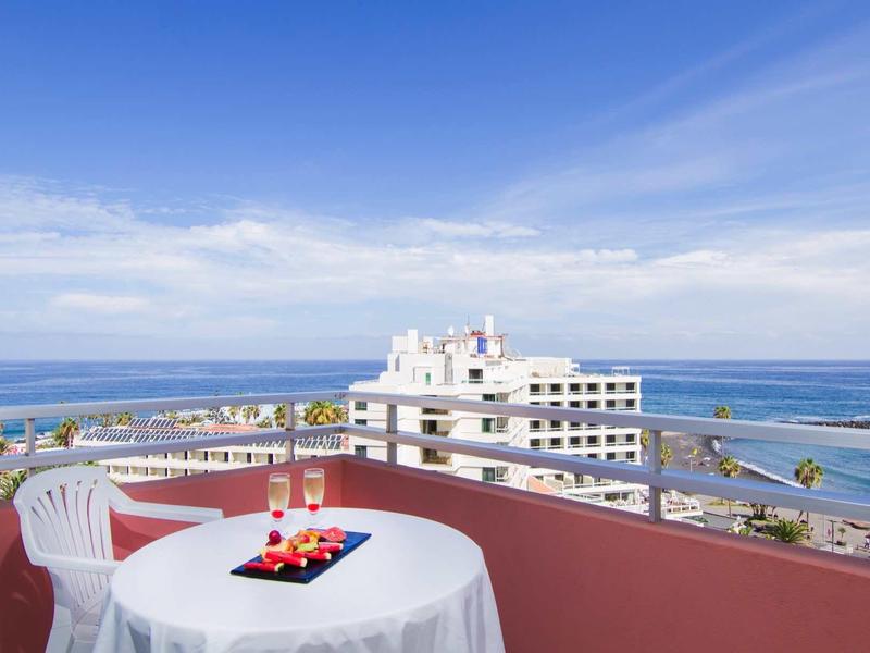 Terrasse mit rundem Tisch, zwei weißen Stühlen, Blick auf Meer und weiße Gebäude unter blauem Himmel.