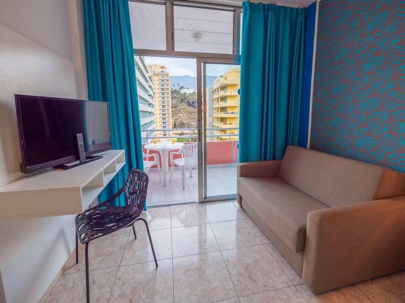 Kleines Wohnzimmer mit braunem Sofa, schwarzem Stuhl, weißem Tisch, TV und blauen Vorhängen vor Balkon.