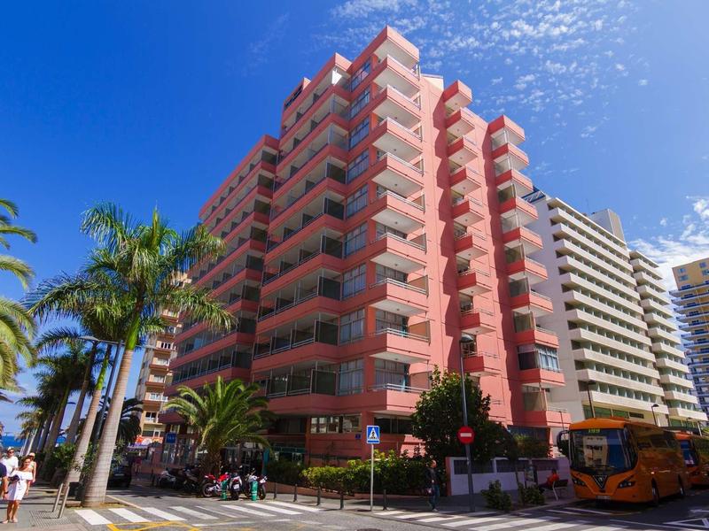 Hohes rotes Wohngebäude mit Balkonen, Palmen und blauem Himmel im sonnigen Stadtbild.