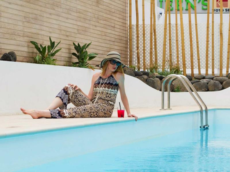 Frau mit Sonnenhut und gemustertem Outfit sitzt entspannt am Beckenrand eines Pools.