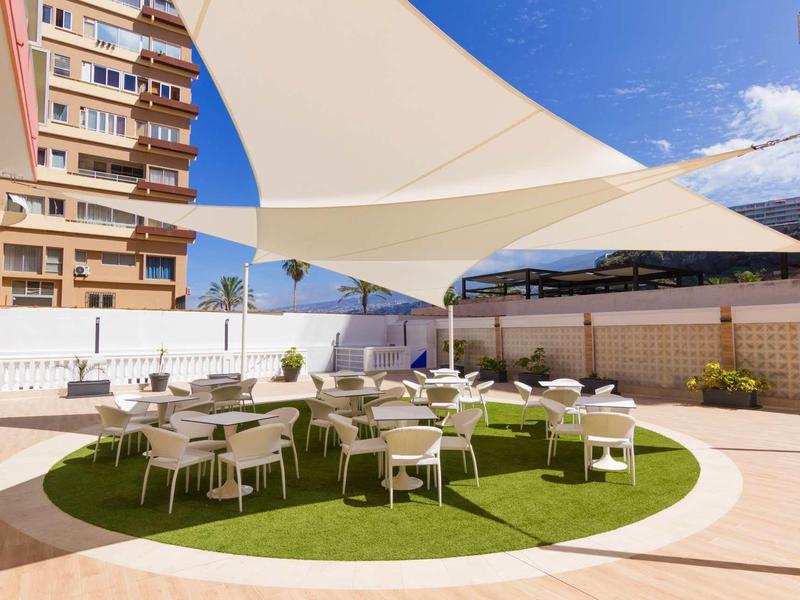 Dachterrasse mit weißen Sonnensegeln, runden Tischen und Stühlen auf grünem Teppich.