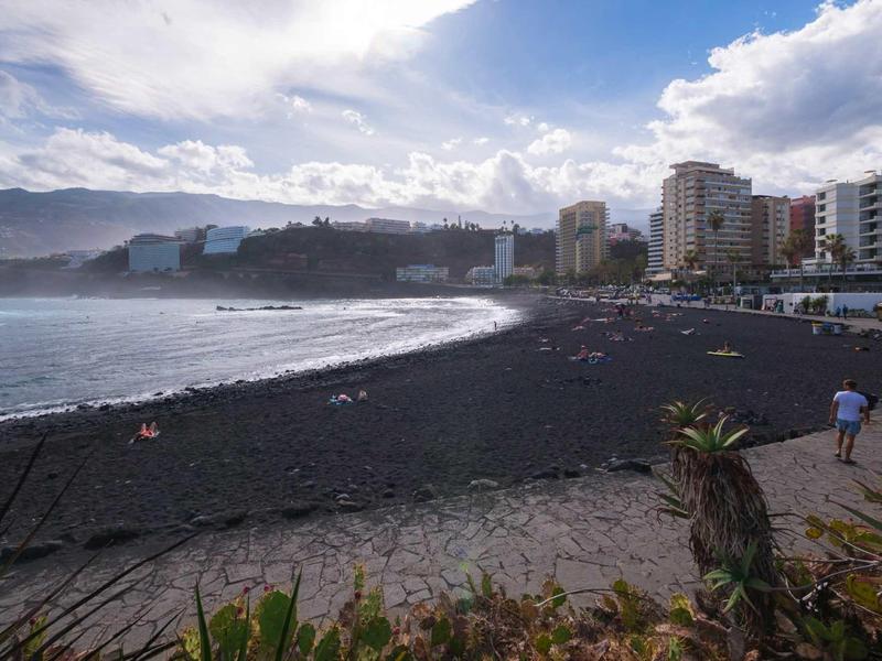Schwarzer Sandstrand an der Küste mit Stadtgebäuden und bewölktem Himmel im Hintergrund.