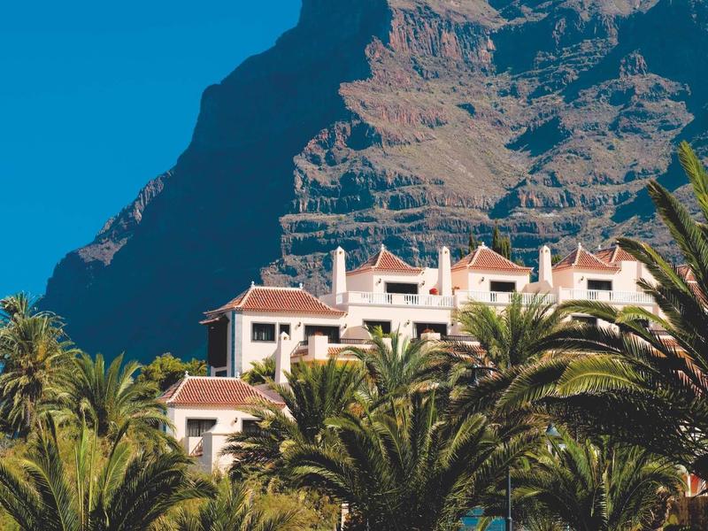 Hotel met rode daken tussen palmbomen voor steile kliffen onder een blauwe lucht.