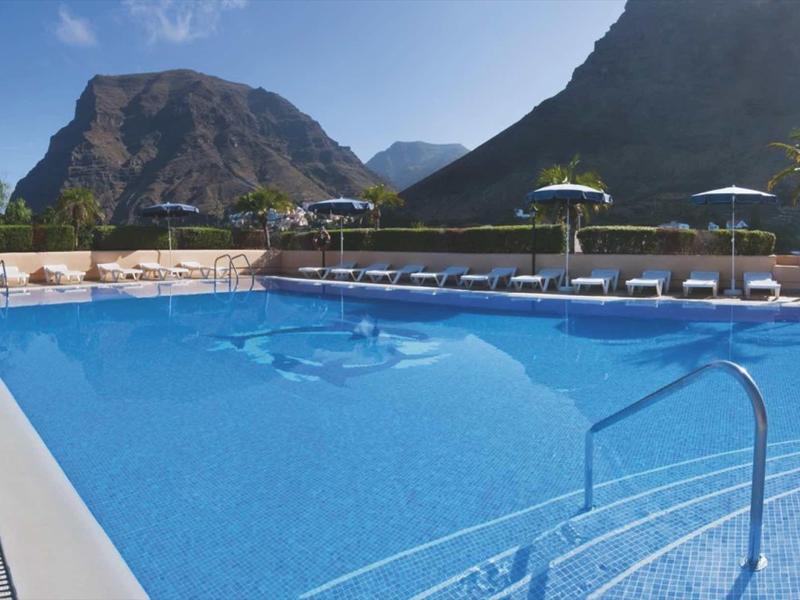 Groot buitenzwembad met ligstoelen en bergen op de achtergrond onder een heldere hemel.