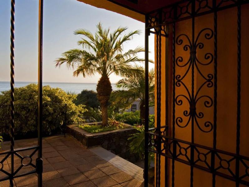 Open deur met smeedijzeren hek, uitzicht op palmbomen en zee in warm avondlicht.