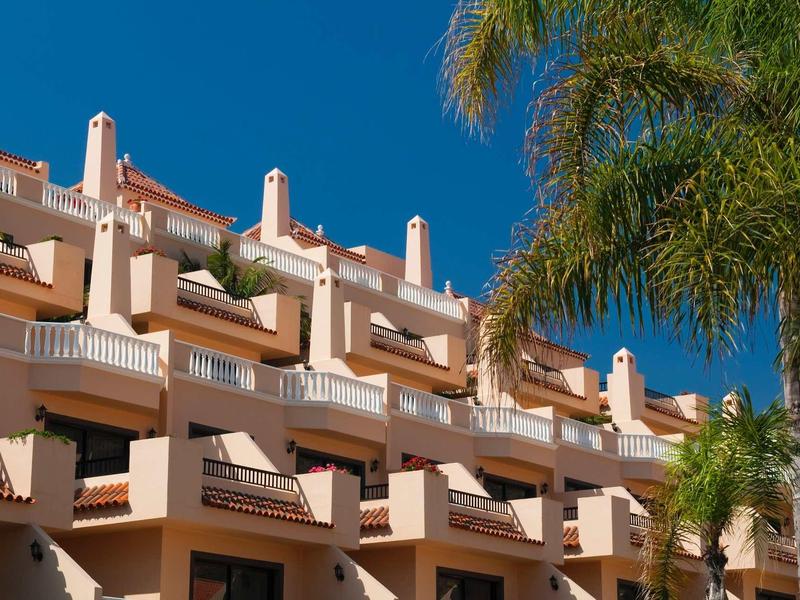 Meerdere verdiepingen hotelgebouw met terrassen en palmbomen onder een heldere blauwe lucht.