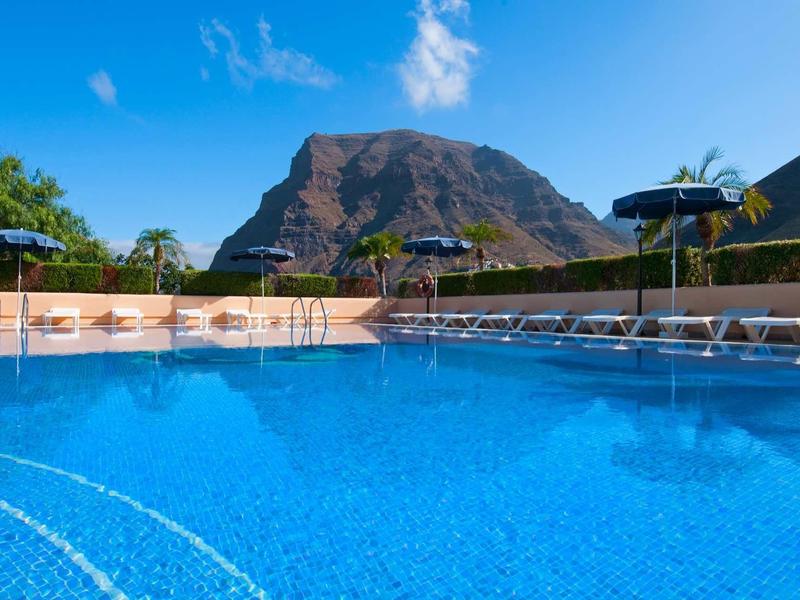 Groot helder zwembad met ligbedden en parasols voor een berg bij een zonnige hemel.