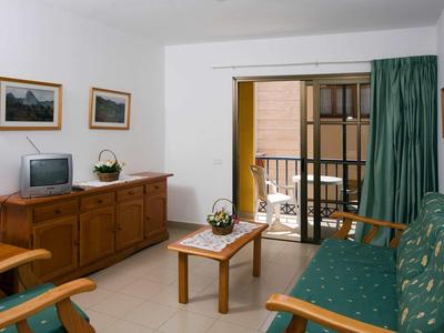 Wohnzimmer mit grün gepolstertem Sofa, Holzmöbeln, kleinen Tischen und Glas-Schiebetür zum Balkon.