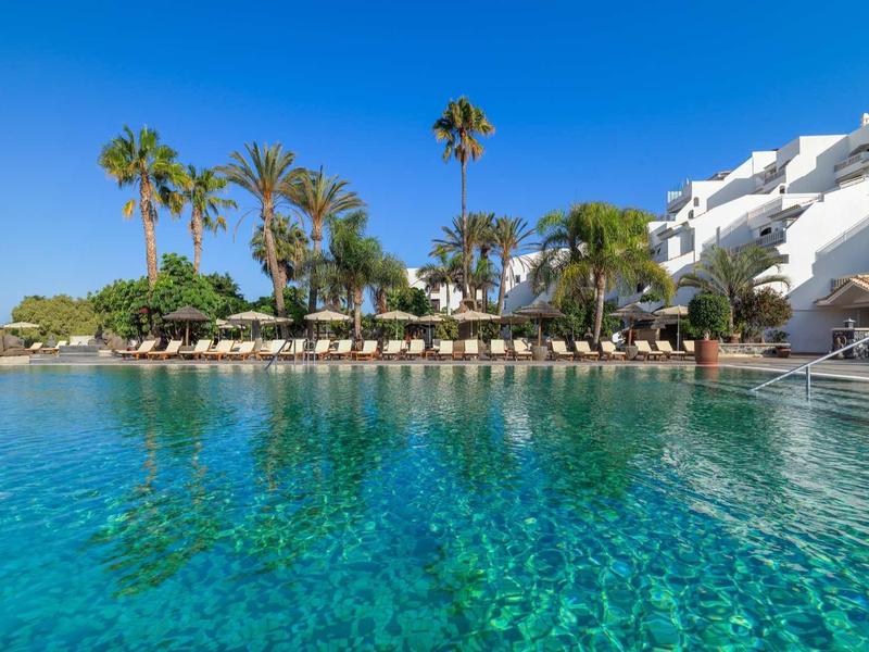 Czysta woda i palmy przed białym budynkiem hotelowym pod słonecznym niebem.