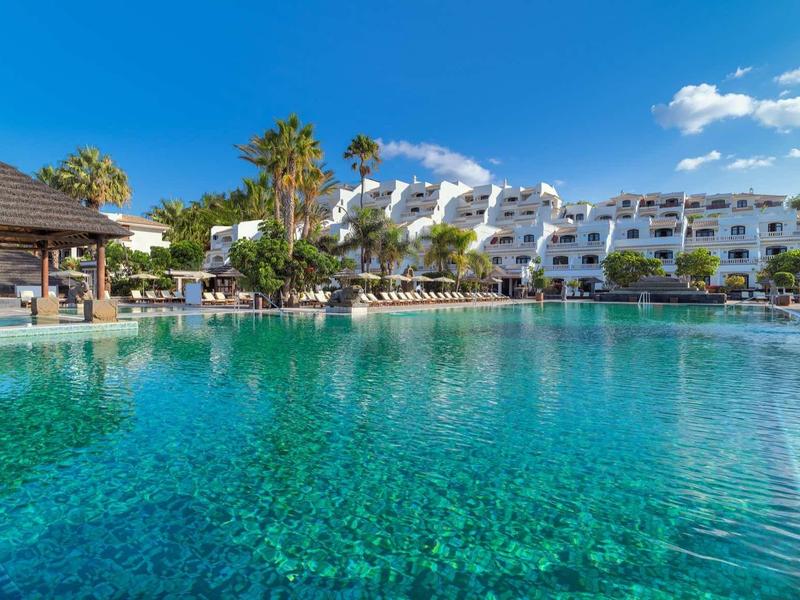 Duży basen hotelowy z czystą wodą, otoczony parasolami i palmami pod niebieskim niebem.