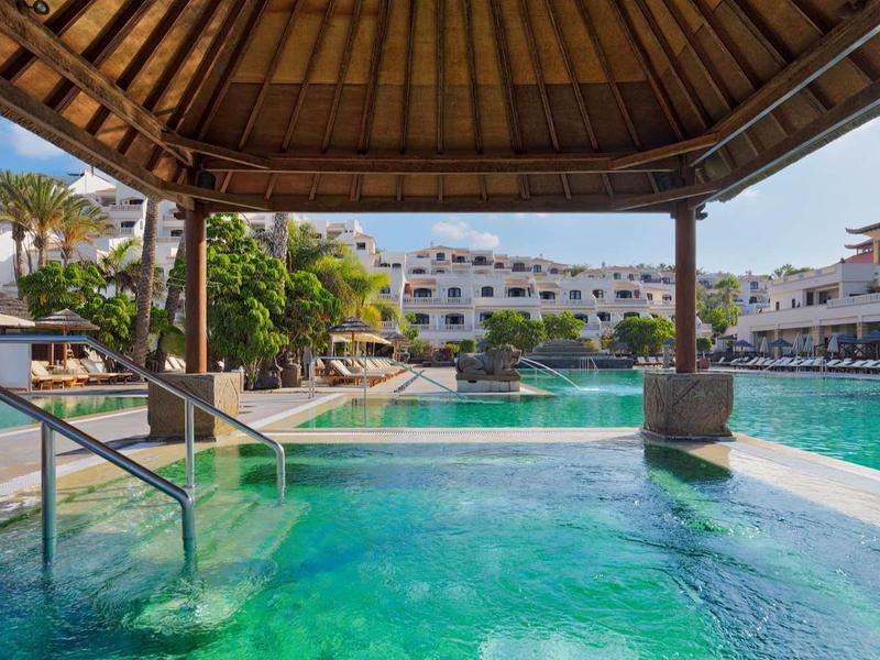 Duży hotelowy basen z zadaszonym jacuzzi i nowoczesną architekturą w tle.