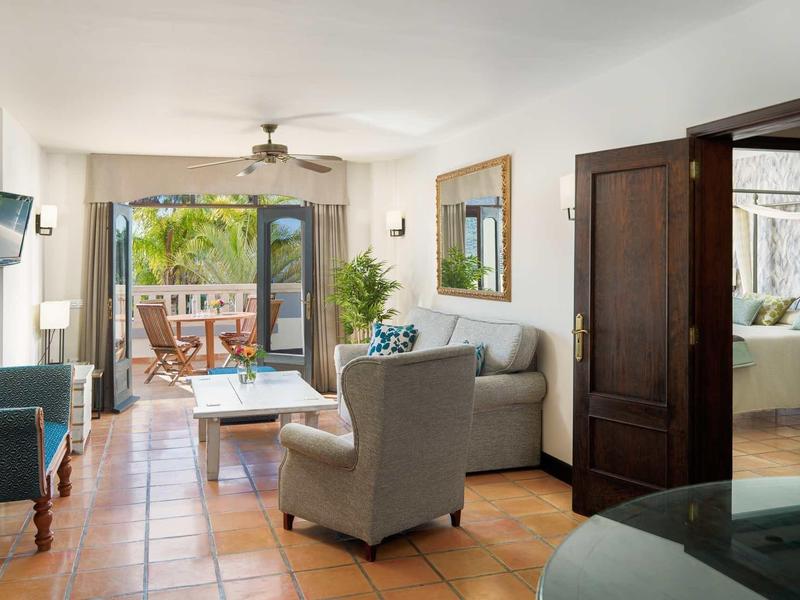 Jasny pokój hotelowy z wygodnym miejscem do siedzenia i balkonem z widokiem na palmy.