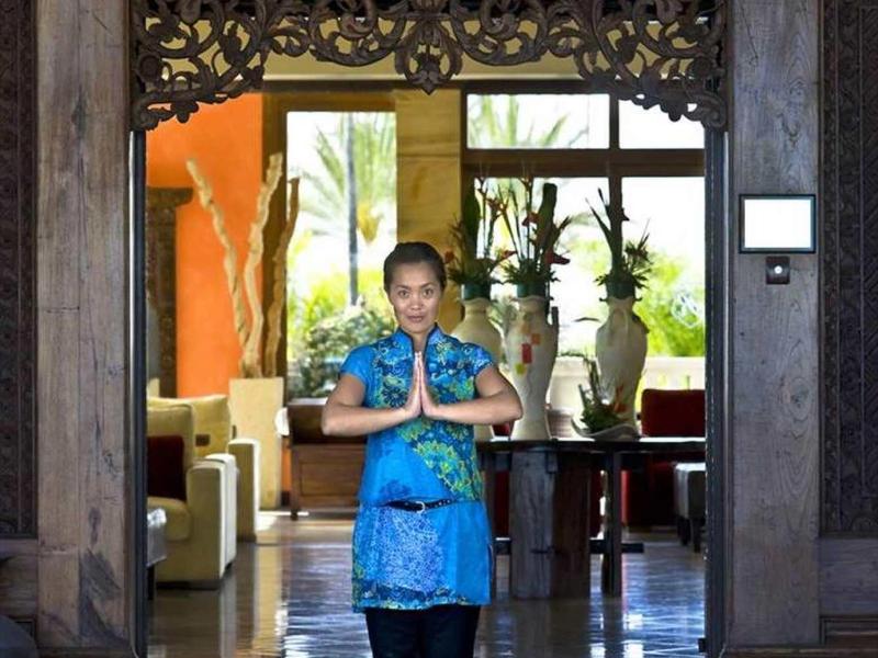 Kobieta w tradycyjnym stroju stoi powitalnie przy wejściu do hotelu z ozdobnymi drewnianymi drzwiami.