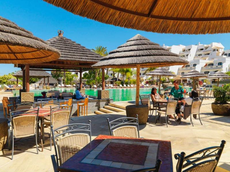 Słoneczny taras na zewnątrz z stołami, krzesłami i parasolami w ośrodku wypoczynkowym.