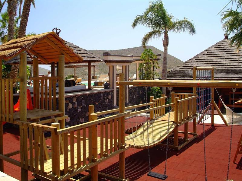 Plac zabaw dla dzieci z drabinką i wiszącym mostem obok hotelowego basenu pod palmami.