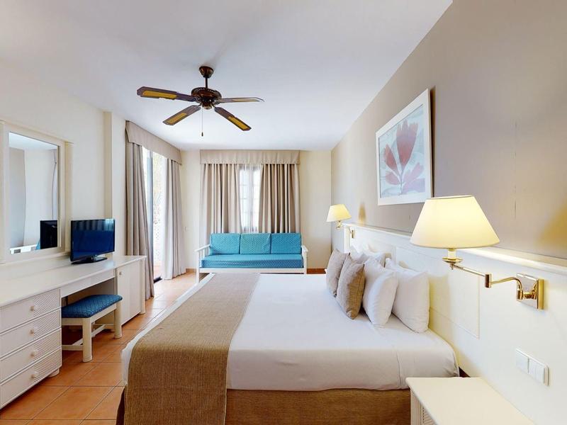 Camera d'hotel moderna con letto matrimoniale, scrivania, TV e area salotto con luce naturale.