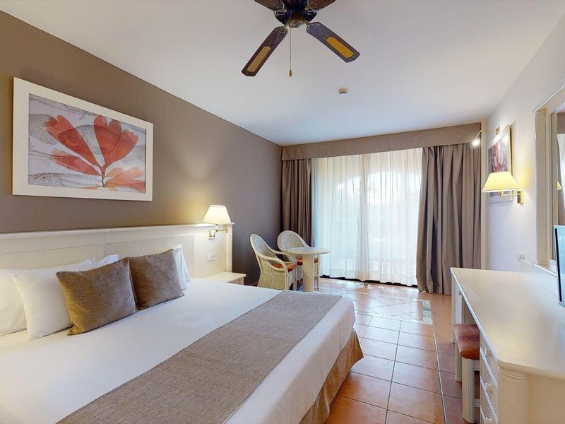 Camera d'hotel luminosa con letto grande, ventilatore a soffitto e scrivania con TV.