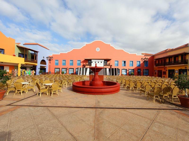 Cortile con edifici rossi, sedie e una fontana rossa al centro sotto un cielo nuvoloso