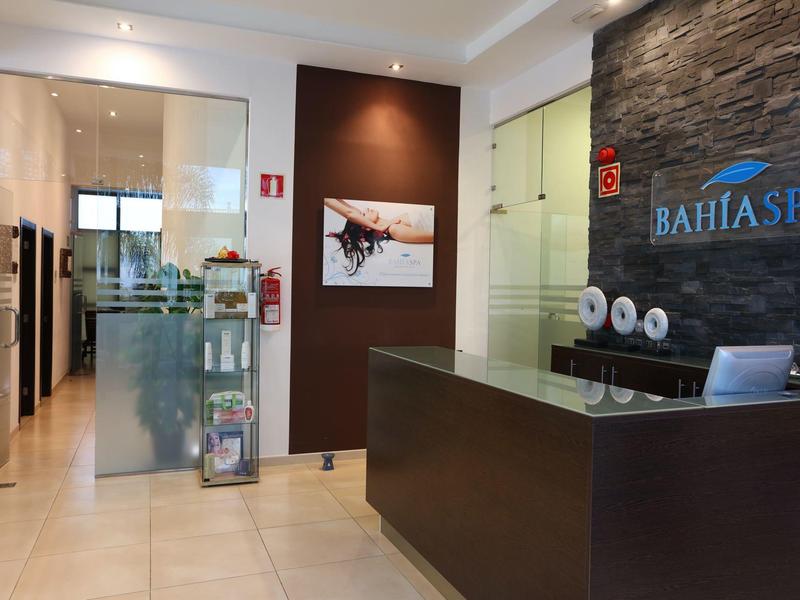 Reception spa luminosa con banco moderno, parete in pietra e corridoio verso l'area trattamenti.