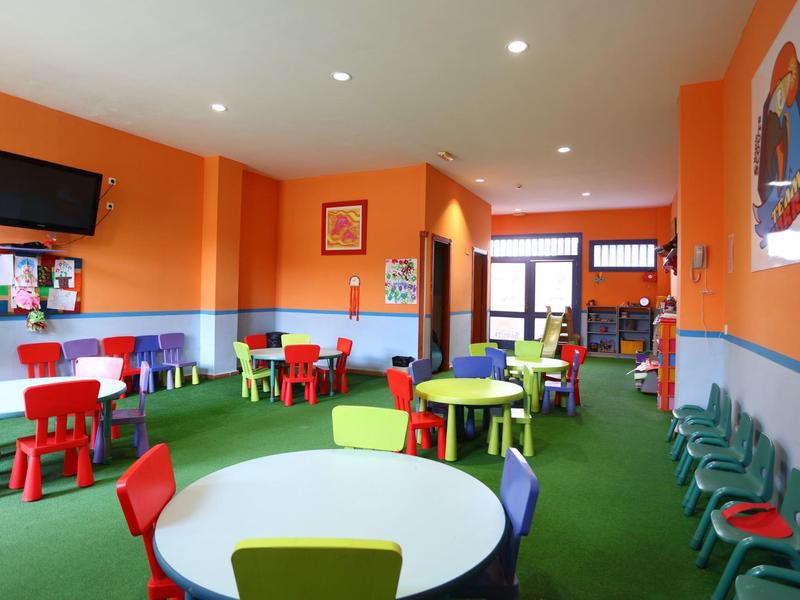 Sala giochi colorata per bambini con tavoli rotondi, sedie multicolori e pareti arancioni.
