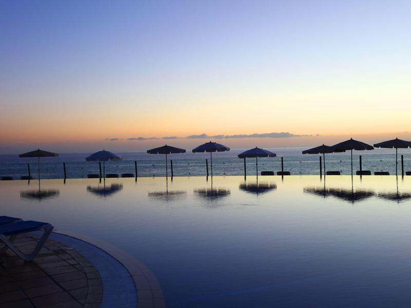Piscina a sfioro con ombrelloni al tramonto accanto al mare calmo.