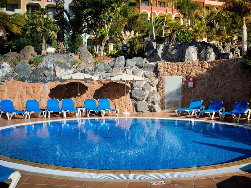 Piscina rotonda con lettini blu e parete rocciosa sullo sfondo in una struttura alberghiera.