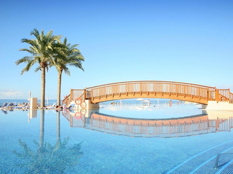Ponte di legno sopra una piscina con palme e cielo azzurro limpido sullo sfondo.