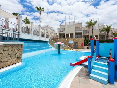 Kinderpool mit blauer Rutsche, Wasserfontäne, Palmen und weißen Gebäuden im Hintergrund bei Sonnenschein.