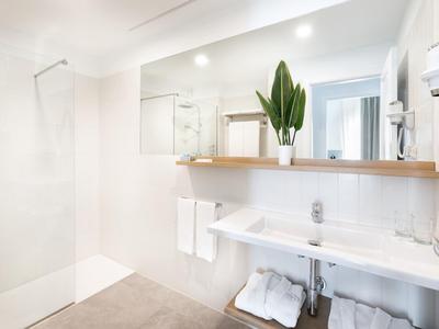 Helles, modernes Badezimmer mit weißem Waschbecken, Regalen, großen Spiegeln und einer Dusche ohne Tür.