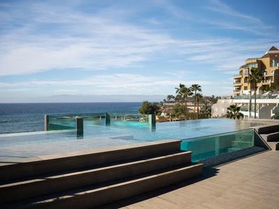 Moderner Infinity-Pool mit Holzstufen, Meerblick und Gebäuden im Hintergrund bei blauem Himmel.