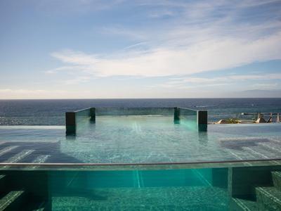 Infinity-Pool mit klarem Wasser, dunkelgrünen Wänden, Blick auf den Ozean und blauem Himmel mit Wolken.