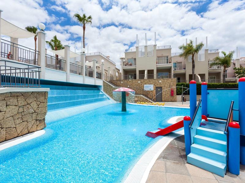 Piscina infantil con tobogán azul, fuente de agua, palmeras y edificios blancos bajo sol.