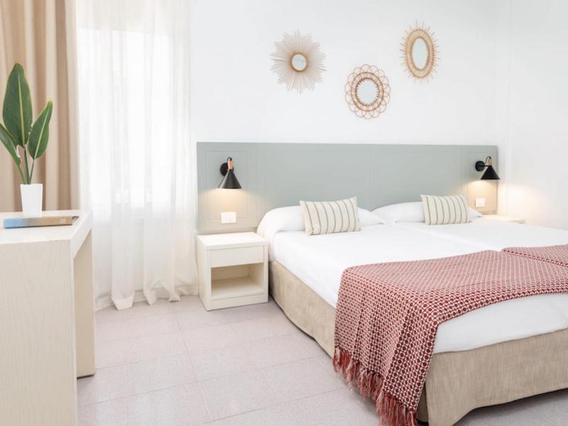 Dormitorio luminoso con cama grande, cabecera gris, mesillas blancas y colcha rosa.