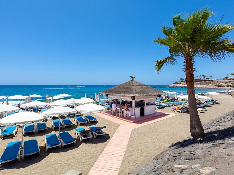 Playa con tumbonas azules, sombrillas, palmera y pequeño kiosco junto al mar bajo cielo azul.