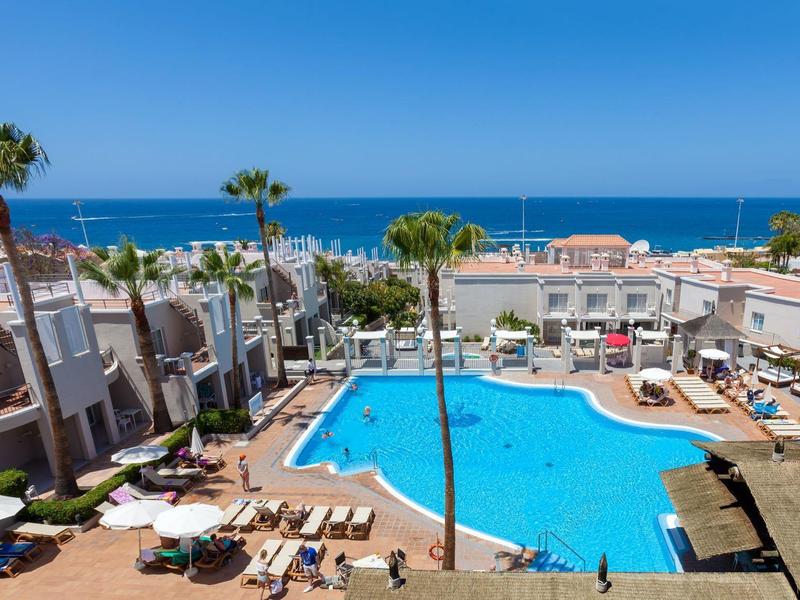 Gran piscina azul rodeada de tumbonas, palmeras y edificios blancos con vista al mar.