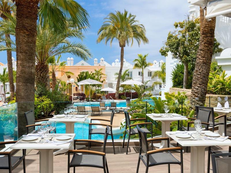 Restaurante al aire libre con mesas preparadas, sillas, piscina y palmeras bajo sol radiante.