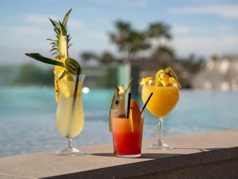 Tres cócteles coloridos con frutas al borde de la piscina bajo cielo soleado y palmeras.