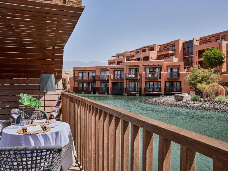 Balkon mit Tisch und Stühlen am Wasser, dahinter moderne rote Gebäude unter klarem Himmel.