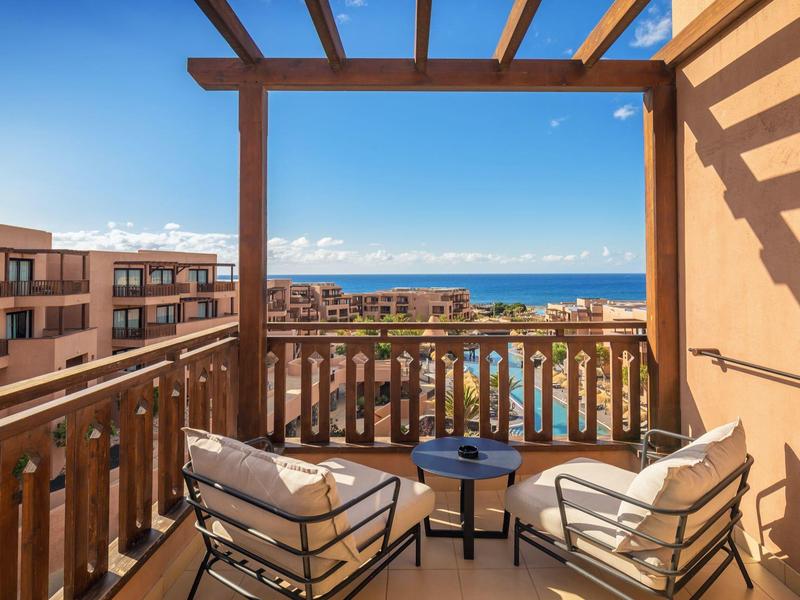 Balkon mit zwei Sesseln, kleinem Tisch und Blick auf Pool, Gebäude und das blaue Meer im Hintergrund.