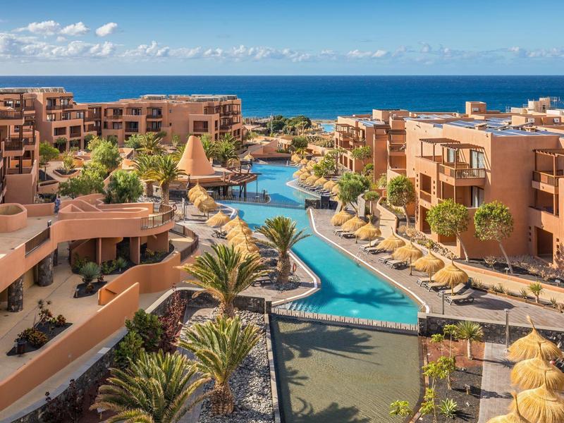 Blick auf ein Hotelresort mit Pool, Palmen, terrakottafarbenen Gebäuden und Meer im Hintergrund.