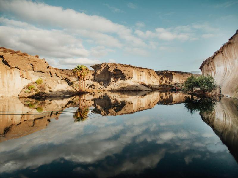 Felsen und Bäume spiegeln sich klar in einem ruhigen See unter blauem Himmel mit Wolken.