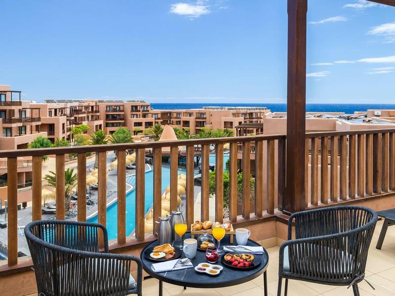 Balkon mit Glasdach, zwei Stühlen und einem Tisch mit Frühstück, Blick auf Pool und Meer.
