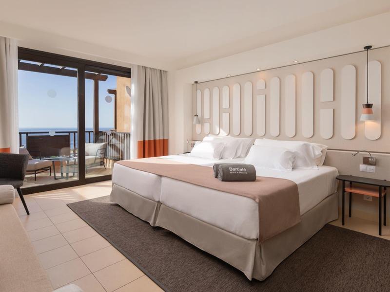 Hell eingerichtetes Hotelzimmer mit Doppelbett, grauem Teppich und Balkon mit Meerblick.