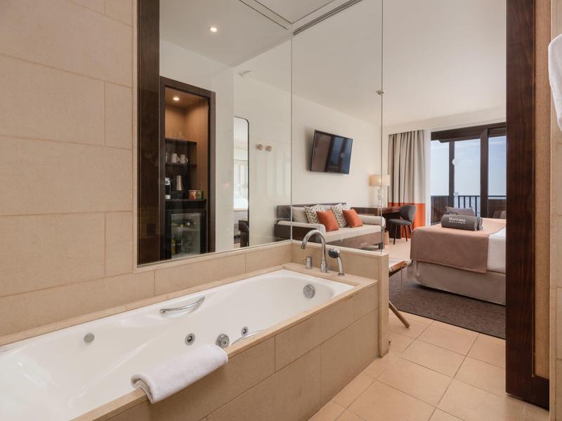 Modernes Badezimmer mit großer weißer Badewanne, Spiegelwand und Blick ins Wohnzimmer mit Sofa.