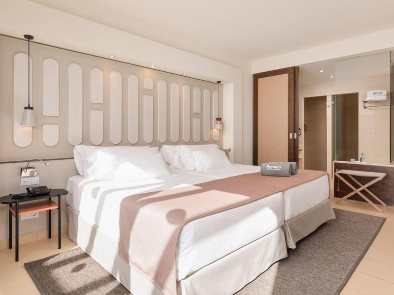 Hell eingerichtetes Schlafzimmer mit Doppelbett, rosa Tagesdecke, Nachttischlampen und großem Spiegel.