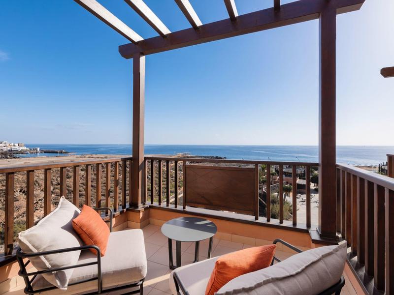 Balkon mit Sitzgruppe, Holzgeländer und Blick auf das blaue Meer bei klarem Himmel.