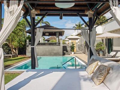 Luxe zwembad met ligbed, witte gordijnen en parasols in een tropische tuinomgeving.